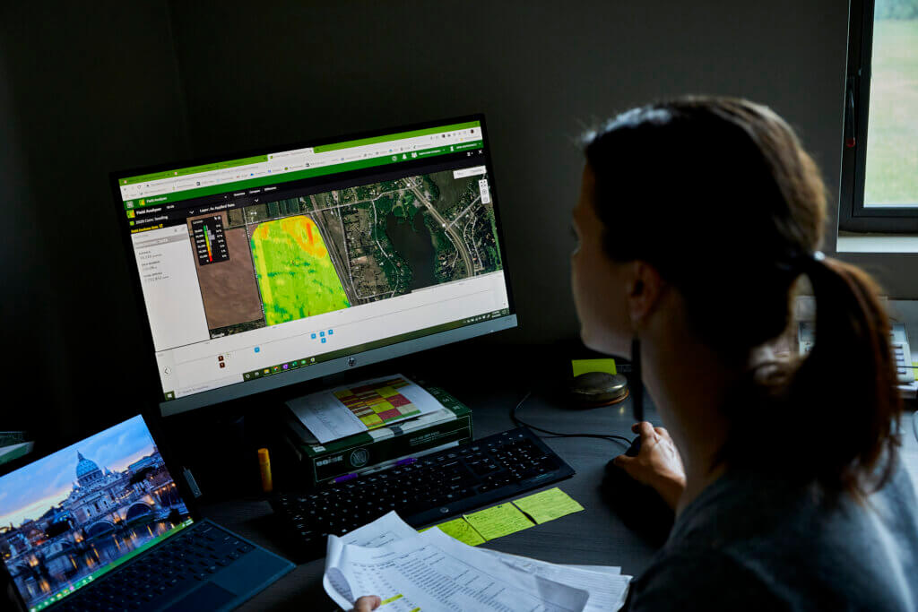 A woman looks at two computer screens, analyzing aerial images of a cornfield to track irrigation management 