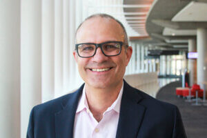 Man with glasses (Marc Hillmyer) poses for a smiling picture in a long contemporary-looking hallway